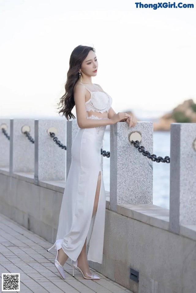 A woman in a white dress leaning against a wall.